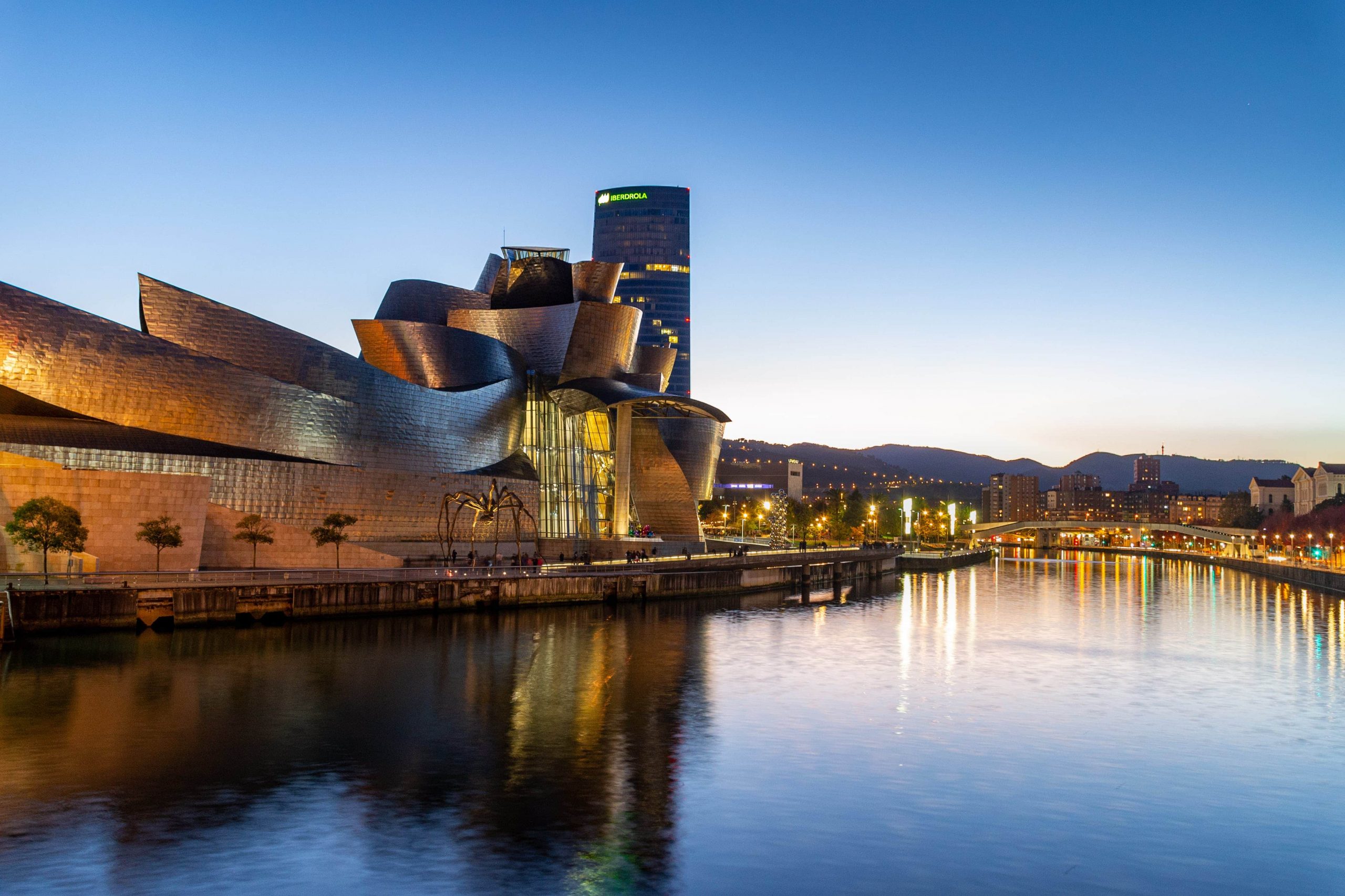 un escena del museo guggenheim de Bilbao al anochecer, con las luces de la ciudad y el propio museo reflejandose en la superficie del agua de la ría de Bilbao